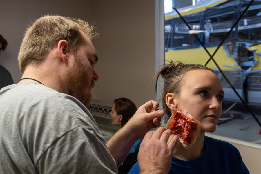 A makeup artist applies a prosthesis and makeup to a volunteer's neck to simulate a realistic looking injury to the flesh