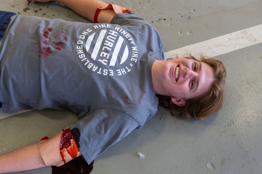 An actor lies on the ground with make-up injuries to his arms and body. He is smiling.