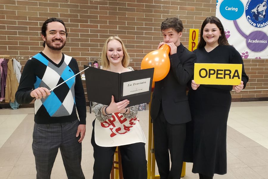 4 ISU music students pose at an elementary school. One is holding a conducting wand, one is holding a musical score, one is blowing up an orange balloon, and one is holding a sign that says 