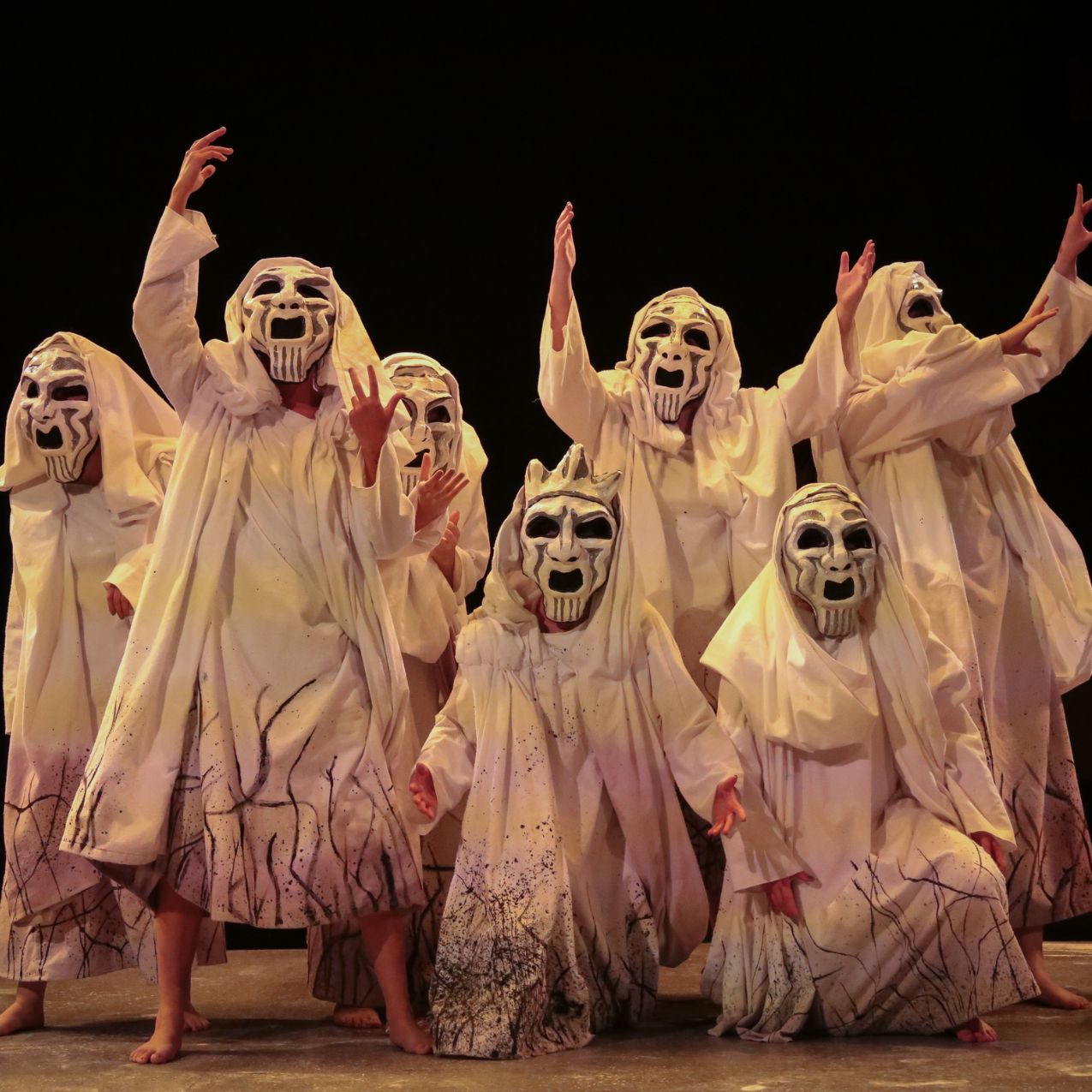 The chorus in the Greek tragedy Burial at Thebes, is onstage. It is dark. The chorus members are all dressed in white rags with white face masks. Their arms are uplifted in a pose of mourning.