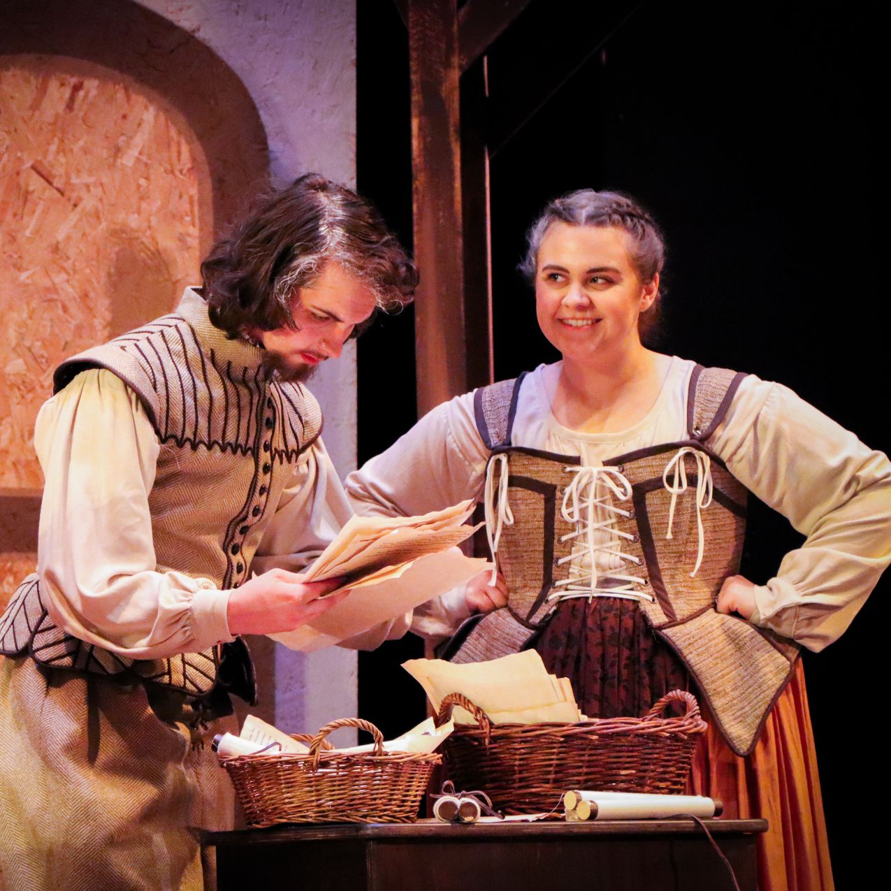 Two actors onstage during a performance of Book of Will: It is a young man and young woman dressed in Elizabethan period clothes, examining a letter on a table.