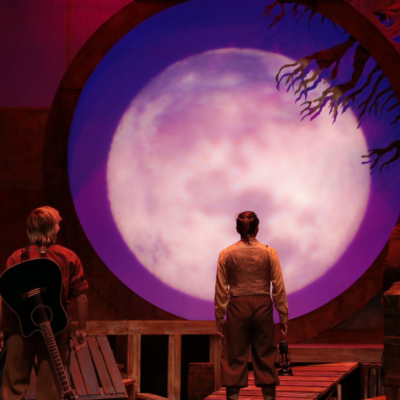 The stage for a performance of The Miraculous Journey of Edward Tulane: The background is lit with a large, rosy moon. Two people face the moon, walking toward it.