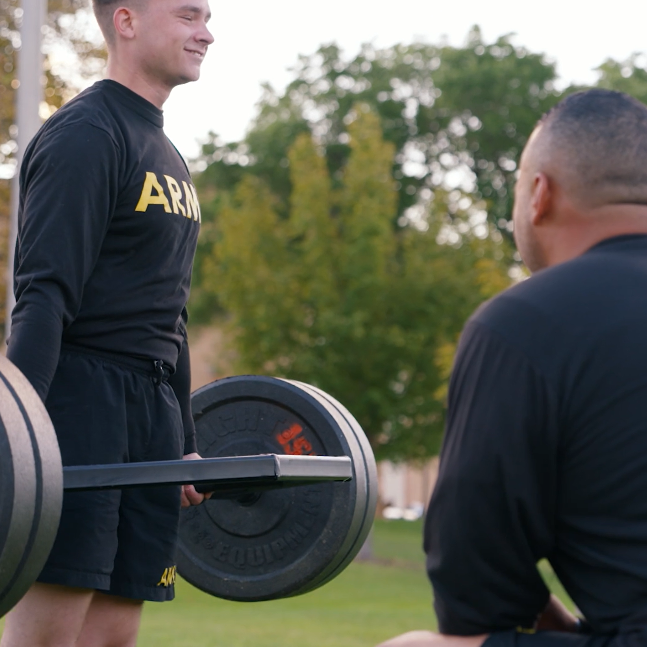 A cadet wearing a black Army sweatshirt deadlifts weights on a barbell. A leader kneels in front of him, looking on.