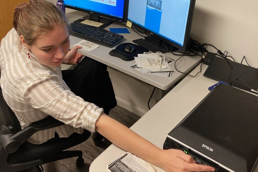 Intern student Katherine Selvage sits at her computer desk and reaches toward a scanning machine.