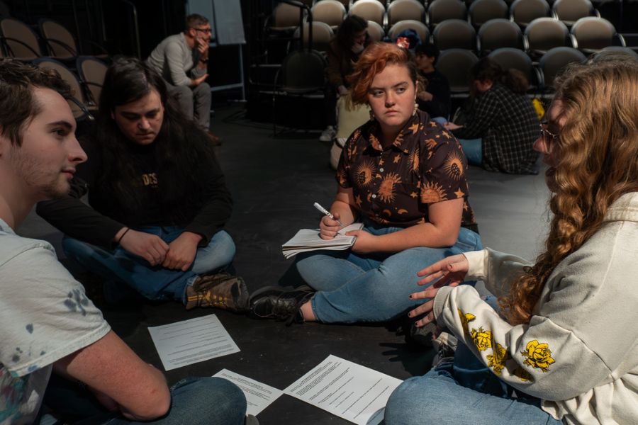 A small group of students sits on the floor of the theatre with notebooks. They are having a deep conversation.