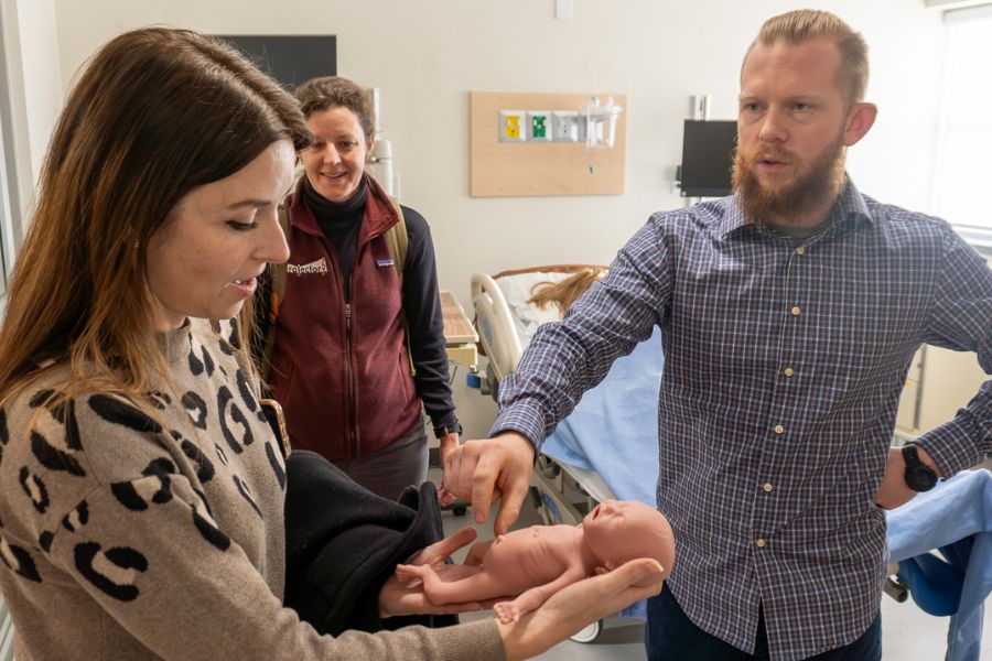 Vanessa Ballam holds a baby dummy while one person looks on and another person points to the baby's chest. They are standing in a simulated hospital room.