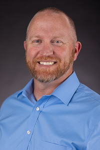 A medium skin toned man with a bald head and red beard. He is wearing a blue button down shirt.