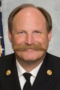 A medium skin toned man with balding brown hair and a large reddish brown mustache. He is dressed in a uniform with a white shirt and a black tie with a suit jacket with two gold buttons. He is smiling