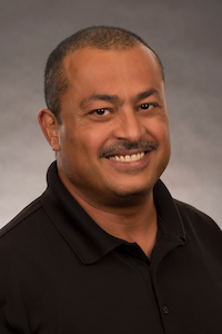 A medium dark skin toned man with a black mustache and hair. He is wearing a black polo shirt and smiling