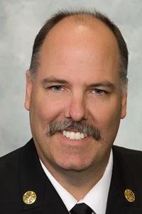 A man with medium skin tone and balding brown hair, and a grey and brown mustache. He is wearing a button down, a tie and a black suit jacket.