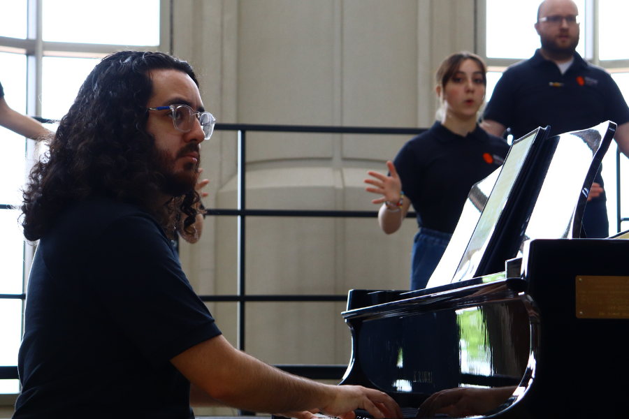 Gabe Lowman sits at a black grand piano in the Stephen's Performing Arts Center Rotunda. Choir members can be seen on risers in the background.