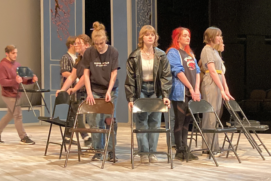 Century High School students act on-stage. They are in normal, modern-day clothing and stand around a ring of metal folding chairs.