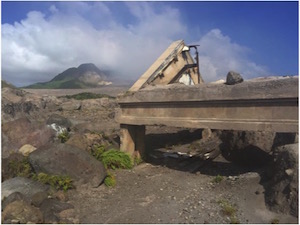 A photo of the devastation caused by the eruption of a volcano on the tiny Caribbean island of Montserrat. Shows were ash has covered the landscape and there is a bridge and road sticking out. 