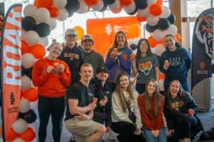 A group of students pose in front of a white, black, and orange balloon arch