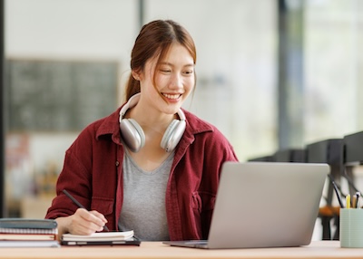 A student sitting behind a laptop with a notepad and pen.