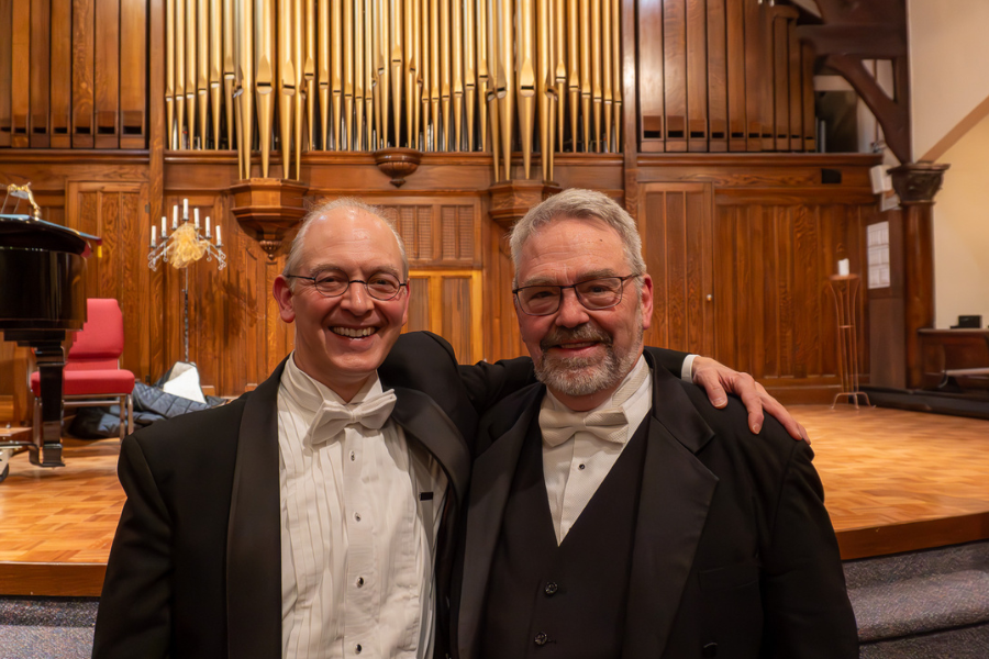 Anderson and Friedley stand with arms around each other and an organ in the background