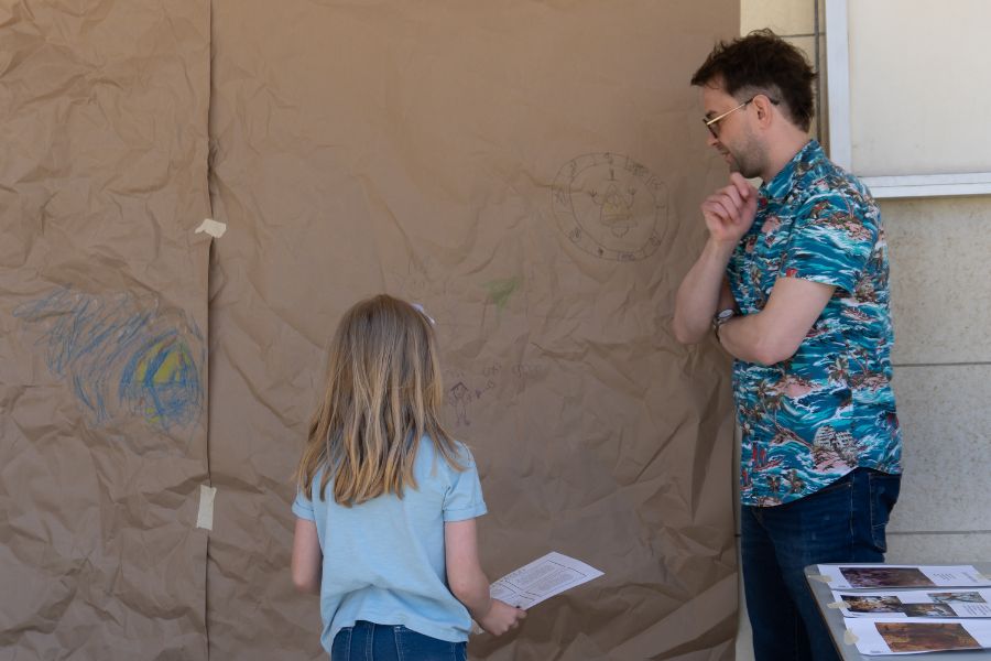 An ISU graduate student watches as a community youth participant plans to color on a big sheet of brown paper attached to the wall