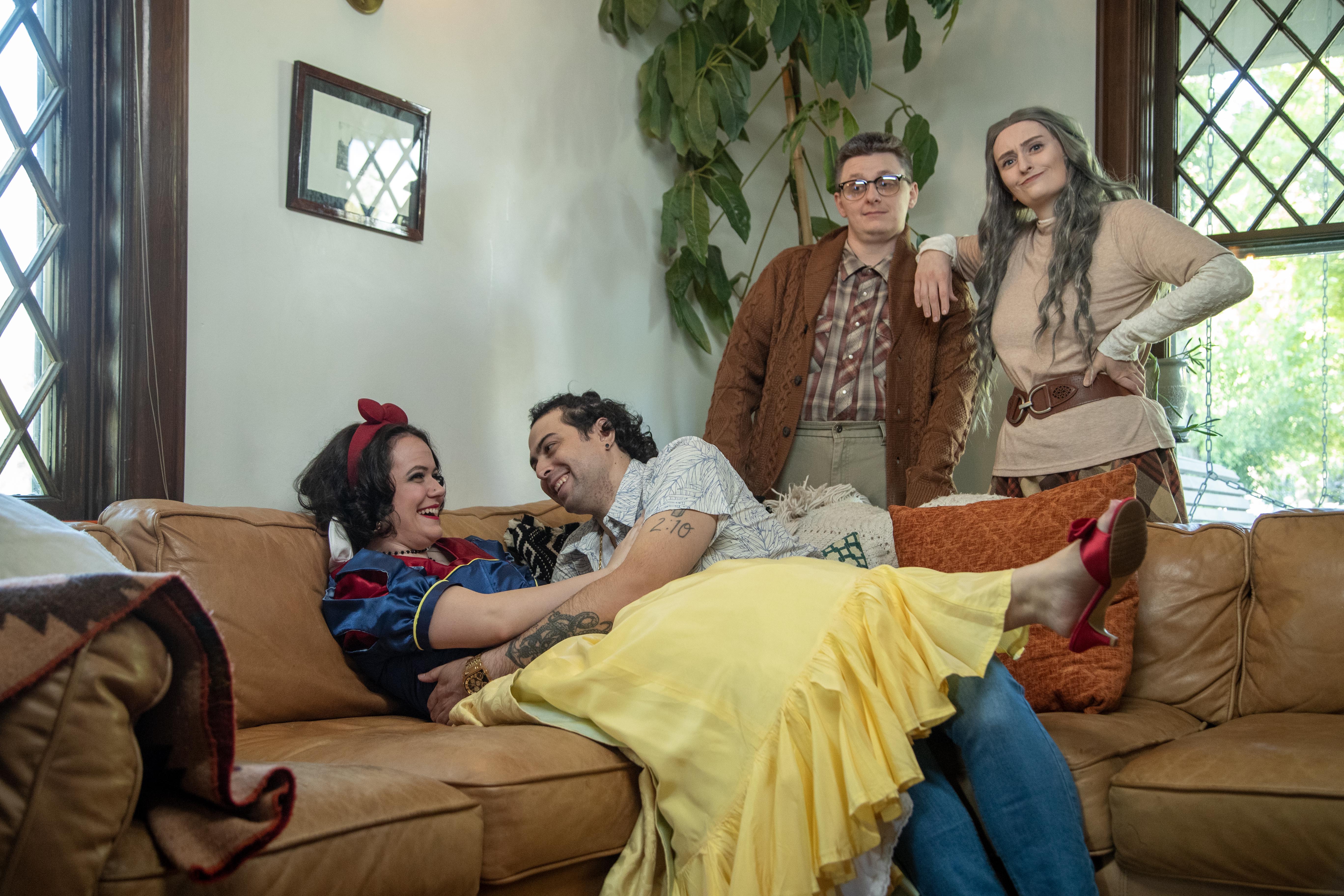 Four people in costume pose in a cozy living room. One couple lounges on a couch, with the woman dressed as Snow White, while two others stand behind them smiling.