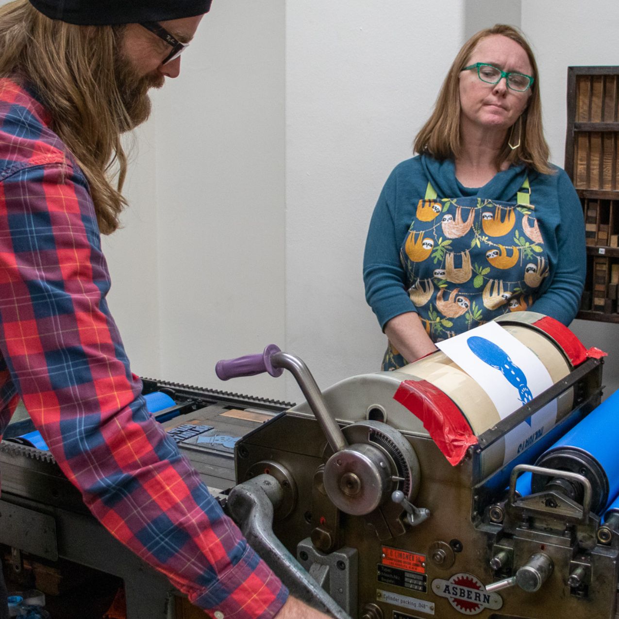 Art faculty member Naomi Velasquez looks on while a student tries working the Pinyon J. Printing Press. He is spinning the handle while the paper is printed with a blue ink. Naomi is wearing an apron.