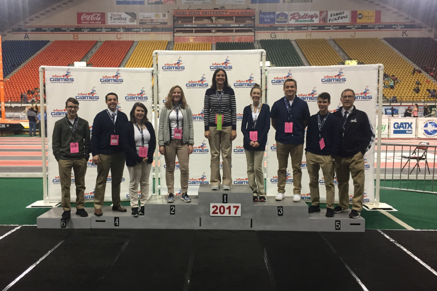 9 people stand on a podium in the Holt arena. They are smiling. There is no crowd in the background.