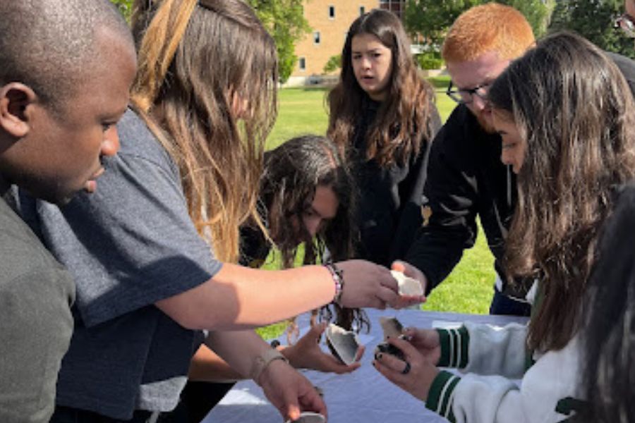 Students gather around a table placed outside on the grass at the quad on the Pocatello campus. They hold pieces of broken pottery and are discussing how to put it back together.