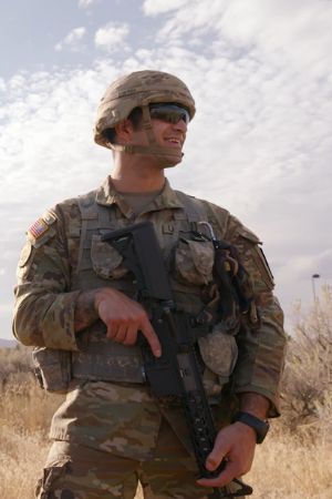 Yousef Elquza stands in a field with a military gun and wearing military camo fatigues and helmet