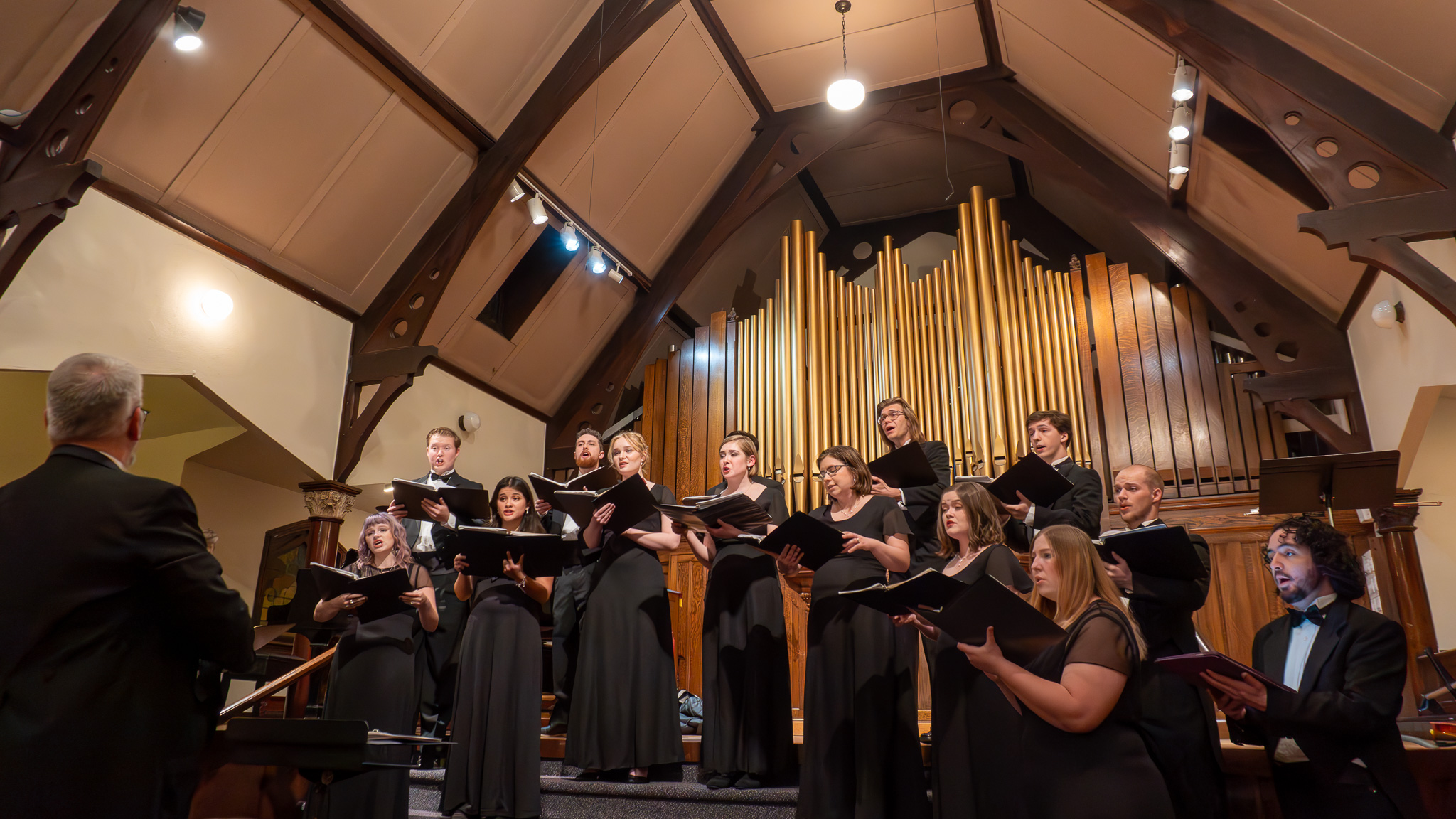 The ISU chamber choir stands with music folders at the front of a church. The organ pipes are behind them and Dr. Anderson directs them.