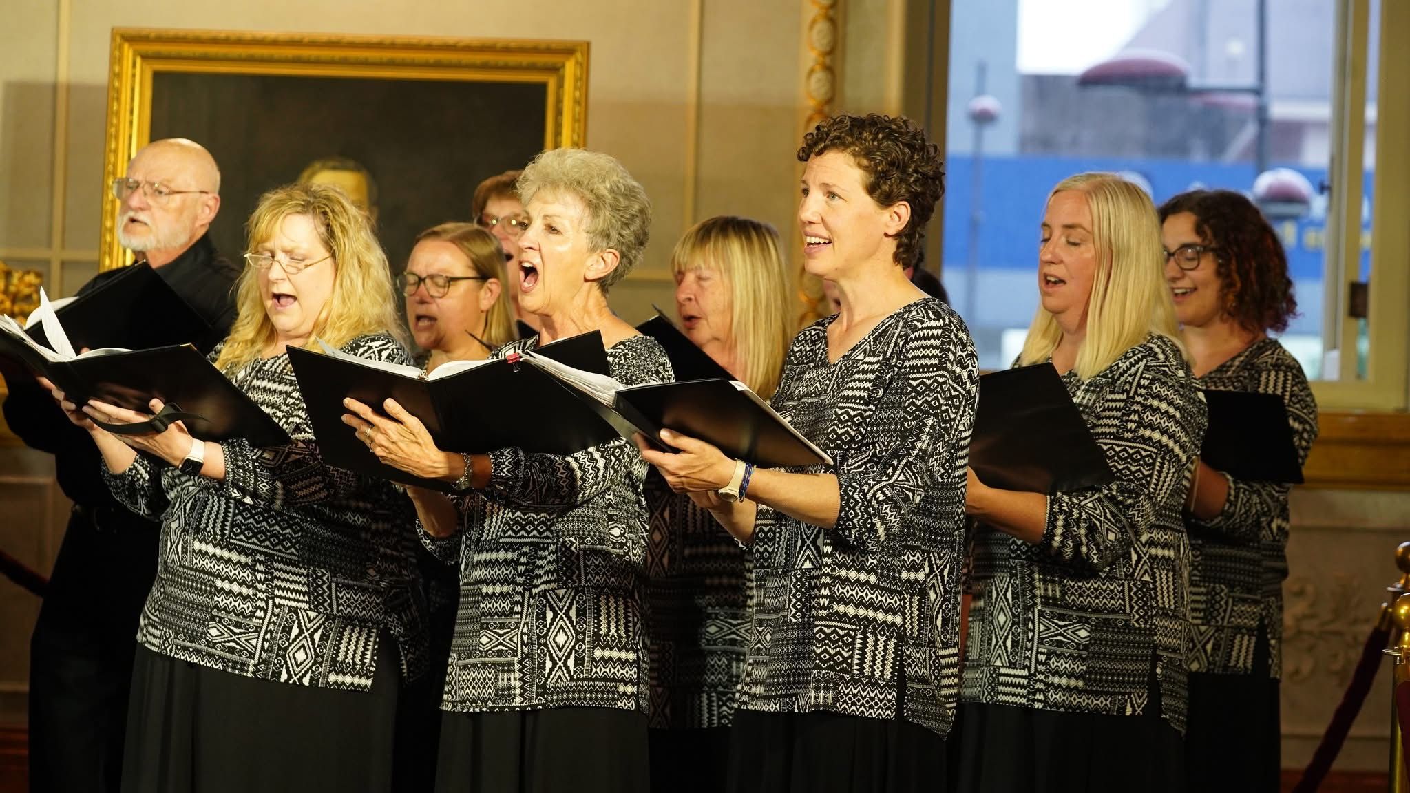 A diverse choir group singing indoors, wearing patterned black and white attire, holding black folders.