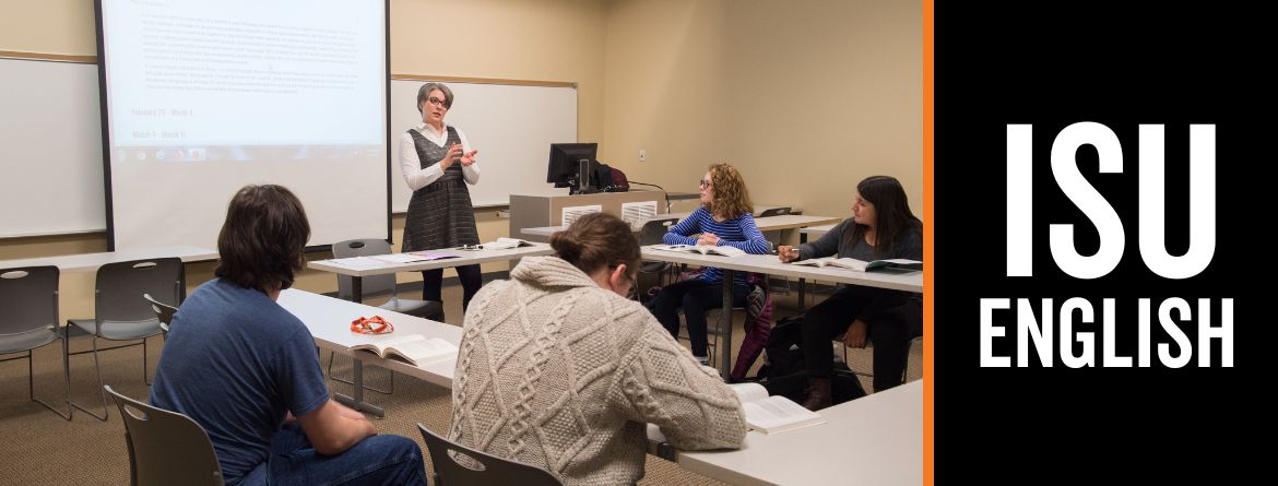In a classroom at ISU, a professor stands at the front of the class in front of a projector screen with text. Students sit at tables in a square formation around her.