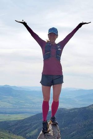 Heather Larson stands on the summit of a hill or mountain with arms upraised to the sky and a big smile.