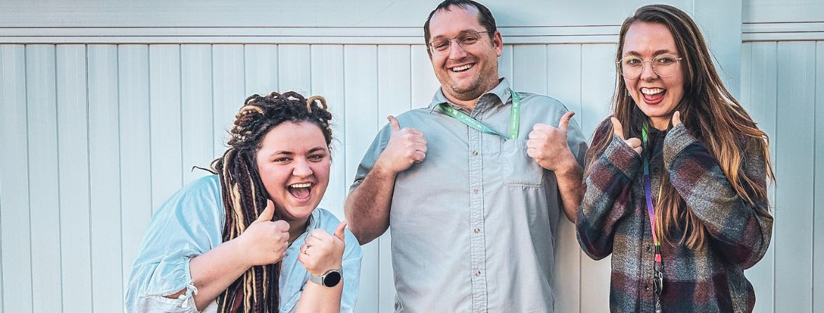 Three people stand in front of a white fence. They are giving the thumbs up sign and have big grins.