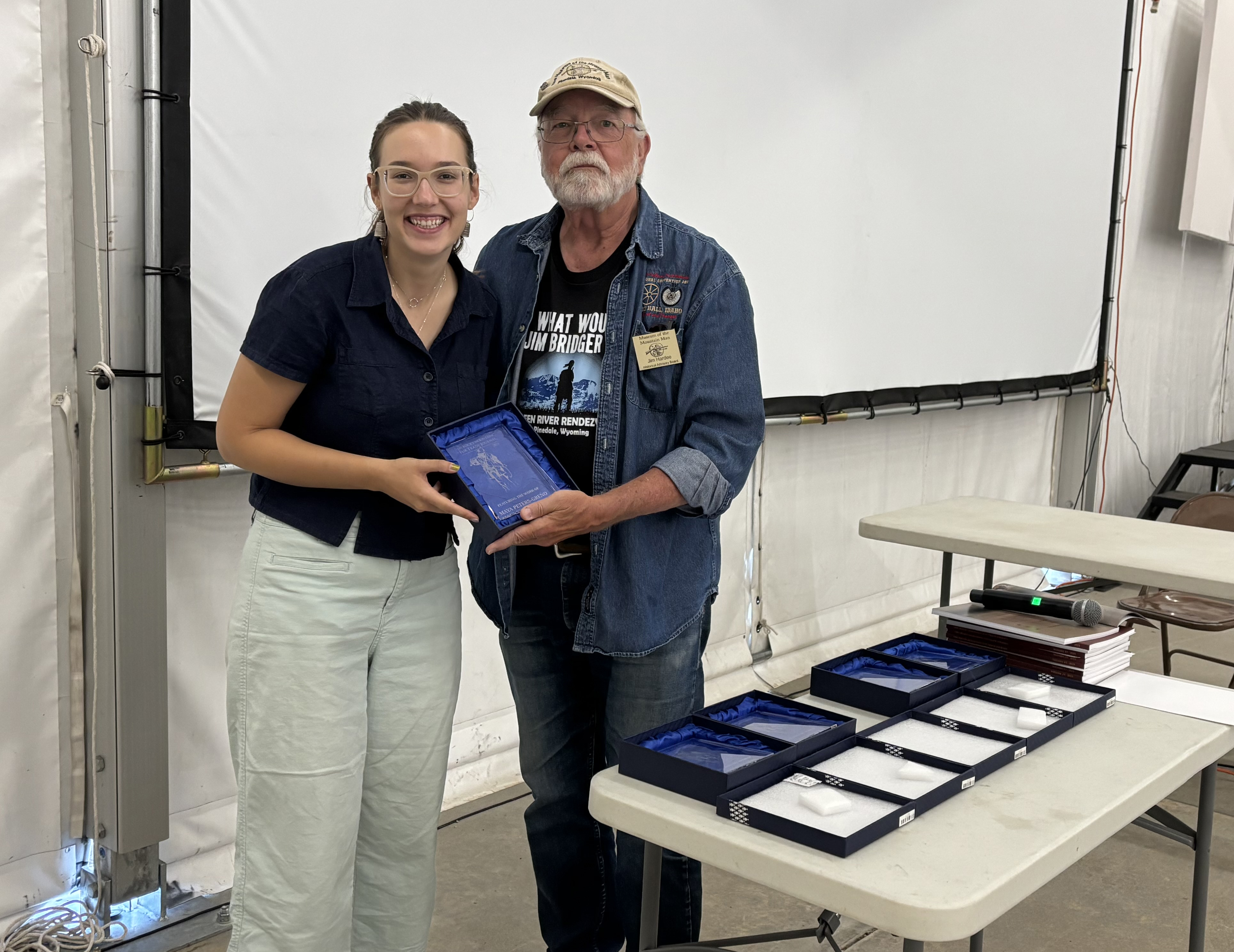 Maya posed with an award plaque in front of a projection screen, with a table of awards
