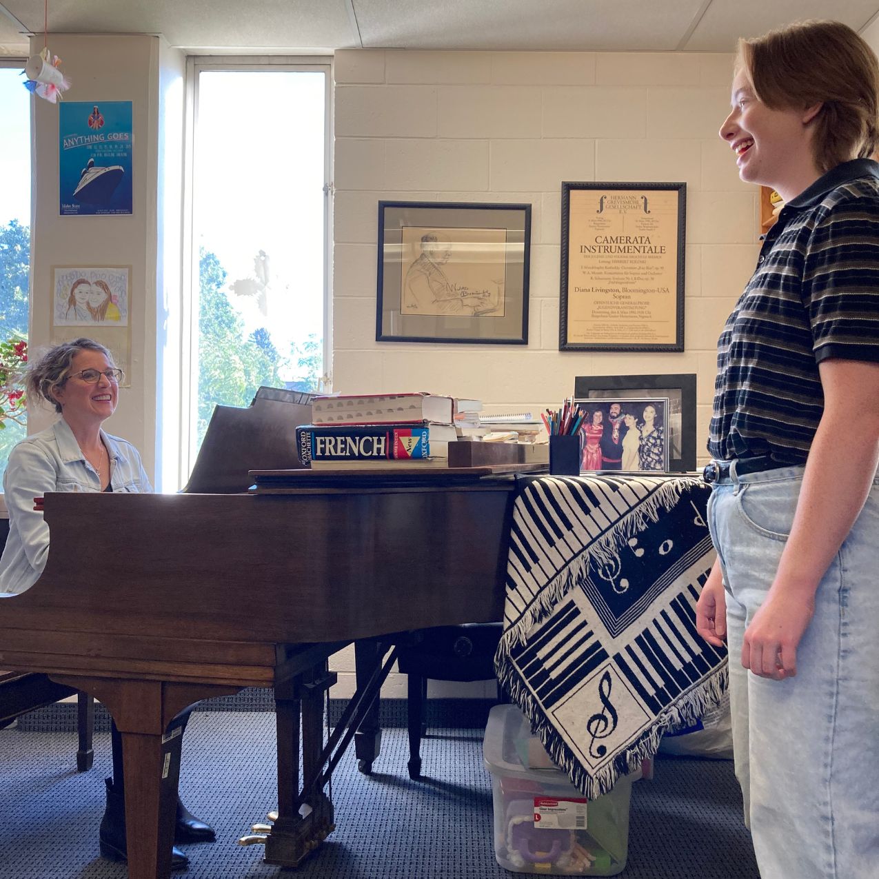 Professor of voice, Diana Livingston Friedley sits at the piano smiling up at her voice student, who is standing next to the piano. They are in a small studio space.
