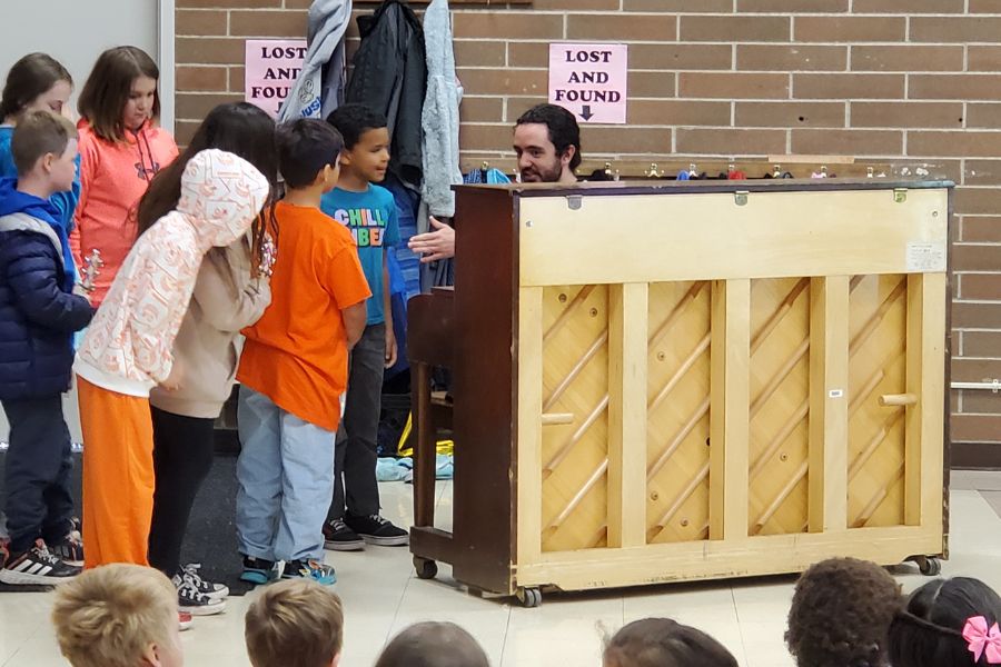 elementary students gather around a piano where Gabe Lowman, ISU alumni is playing the piano