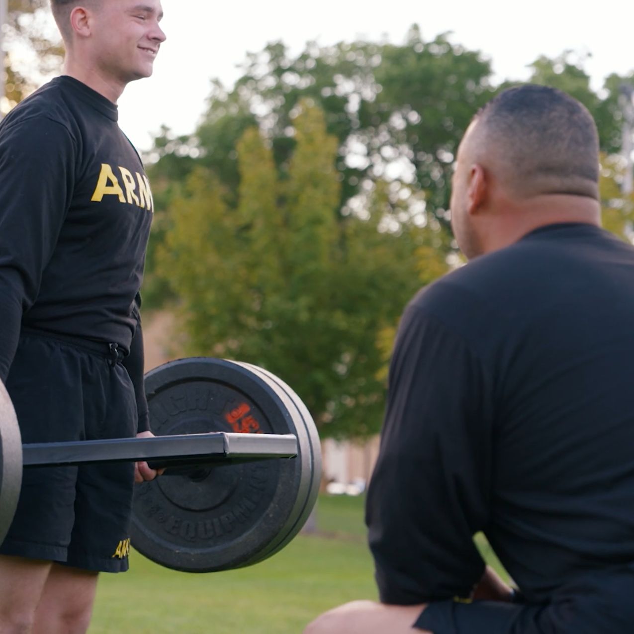 ROTC student cadet deadlisfts a barbell with weight plates on it while his officer looks on. They are outdoors on the grass.