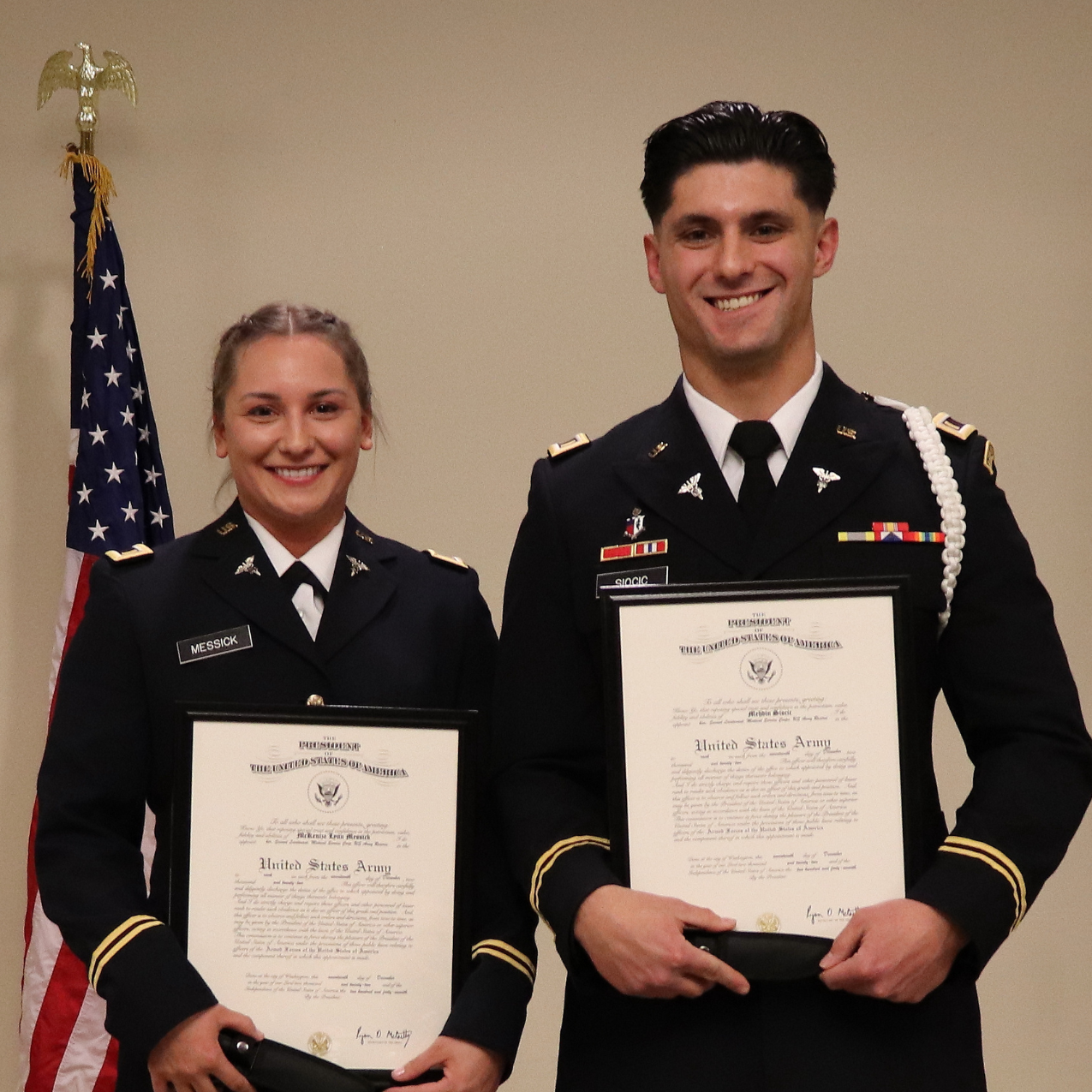 Two graduating cadets, a man and a woman, stand near the US flag in dress uniform, holding their certificates and smiling