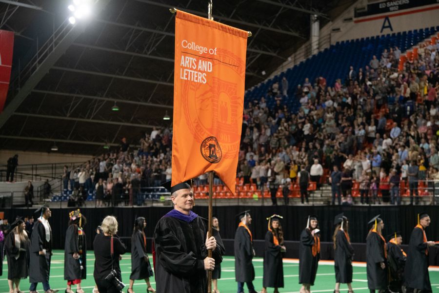 Photo of commencement in the Holt Arena. A line of students in cap and gown follow behind the flag bearer, who carries a banner that says 