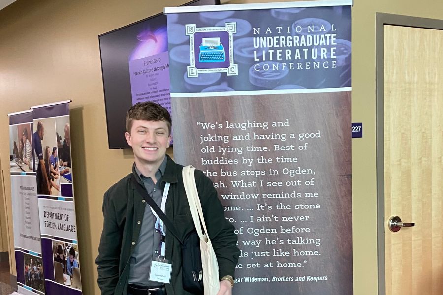 Tanner Pratt stands in front of a banner for the literature conference he is attending. He is smiling.