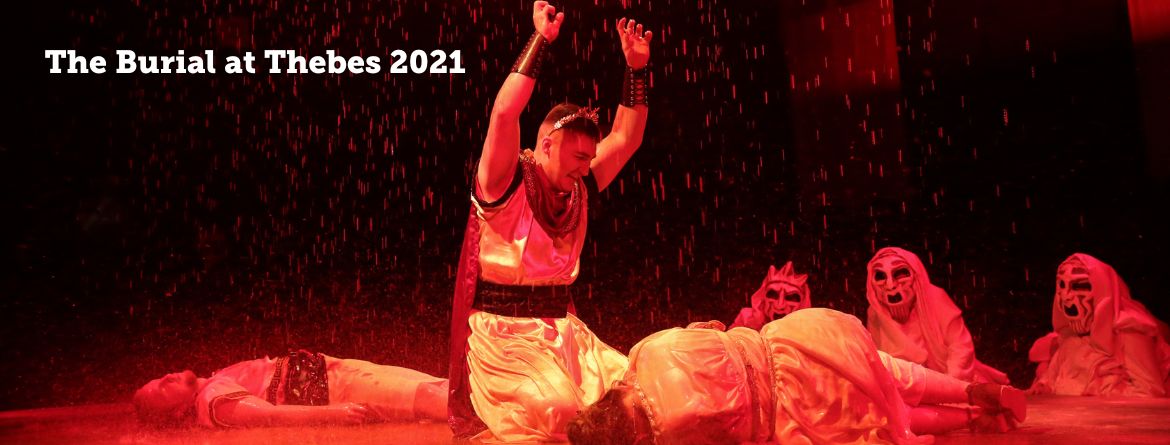 Burial at Thebes 2021. Greek actor kneels next to dead women with fists raised in air in grief while red blood rain falls down