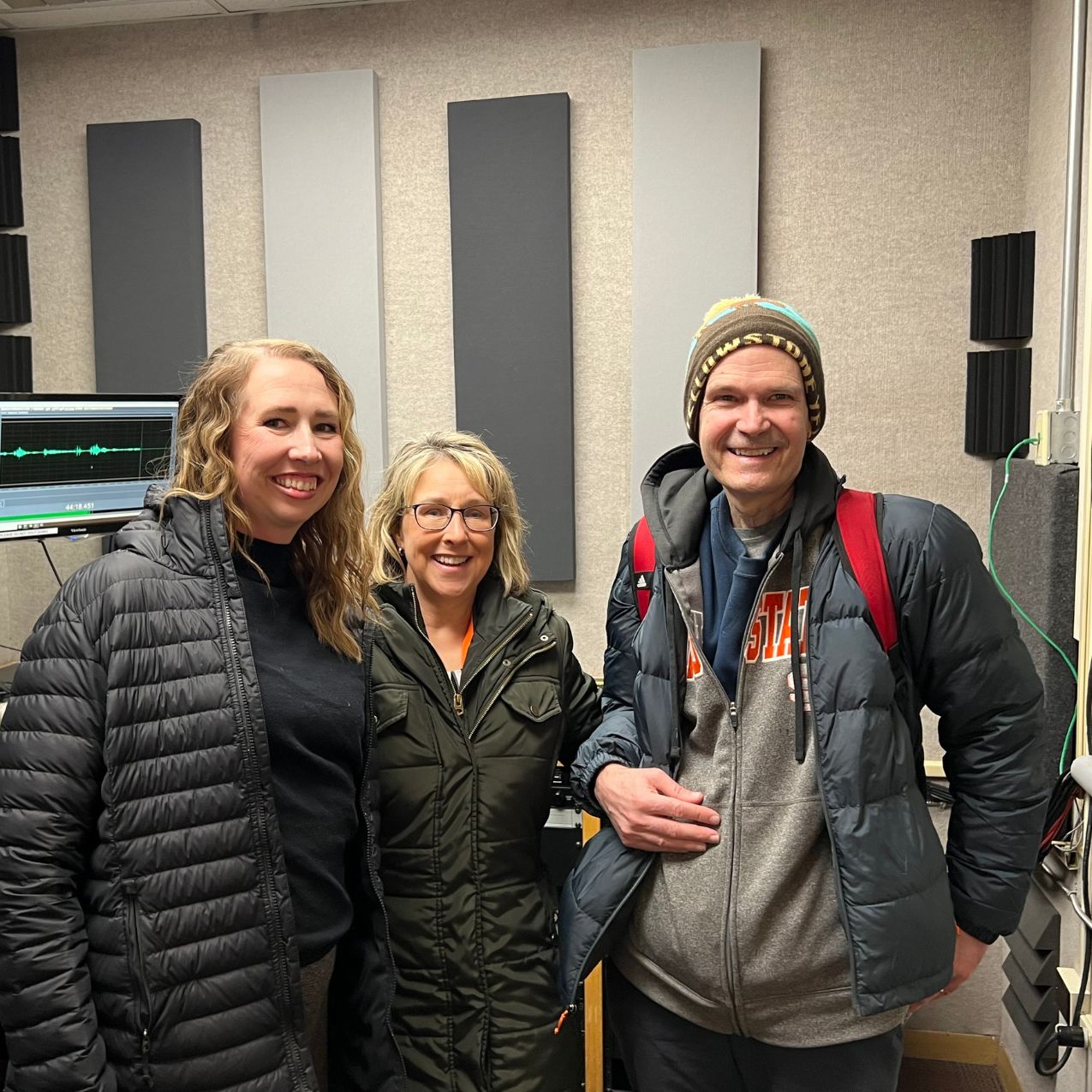 Whitney Fenwich, ISU alumna, Kandi Turley Ames, Dean of the College of Arts and Letters, And Mark McBeth, Proffessor and Chair of the Department of Political Science pose together smiling, in the recording studio. Recording equipment such as a computer screen and speakers are behind them.