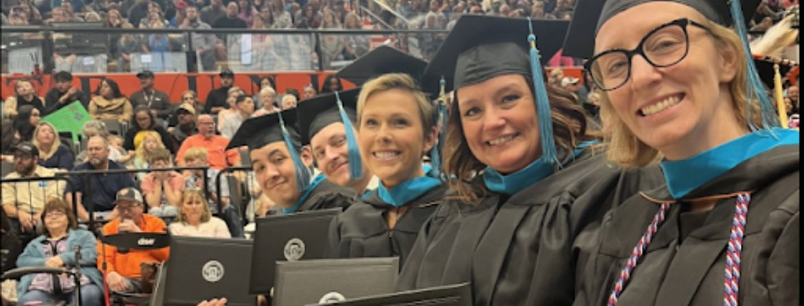 Students at commencement in cap and gown sit in a row holding up their diplomas and smiling