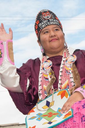 Yvonne Warjack raises one hand in greeting. She is outside in traditional tribal clothing.