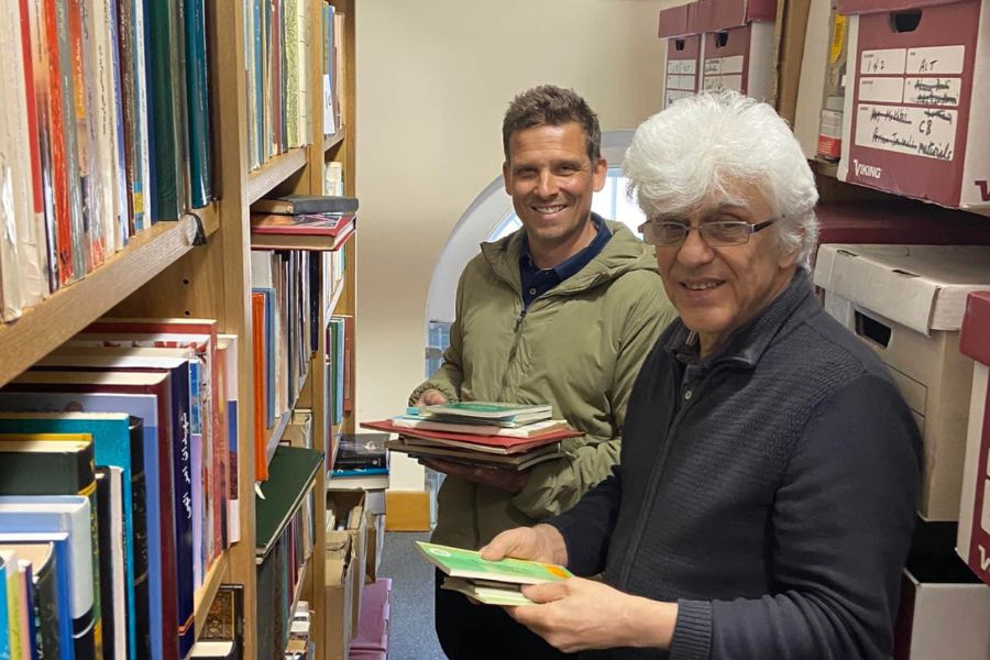 Zack Heern smiles at the camera. He is standing next to another gentleman between bookshelves. They are each holding books.