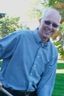 A light skin toned man with balding white hair wearing sunglasses, a blue collared shirt and black pants with a black belt. He is posing with a shovel and smiling. There are trees and grass in the background