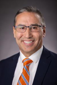 A medium skin toned man with brown hair and glasses. He is wearing a white shirt, red and blue tie, and a black suit jacket.