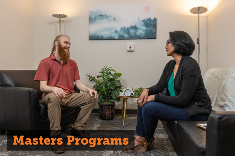 Image: A red-haired man and a dark-haired woman sitting on opposite couches, speaking with engaged body language. Text: Masters Programs. Links to masters programs menu page.