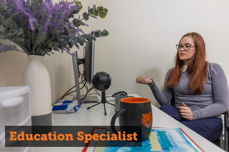 A redheaded woman sitting at a desk facing a computer. Text reads: Education Specialist. Links to education specialist page.