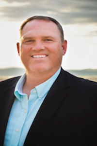 A man with light skin tone and brown hair wearing a light blue button up shirt and a black suit jacket. He is smiling and behind him appear to be mountains.