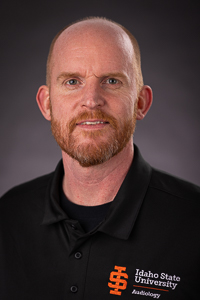 A man with light skin tone with balding red hair and a red beard and mustache. He is wearing a black polo that reads Idaho State University Audiology. He is smiling.