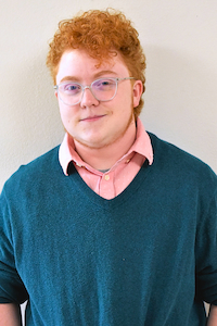 A man with light skin, curly red hair, and blue glasses wearing a pink button up underneath a teal sweater. He is smiling